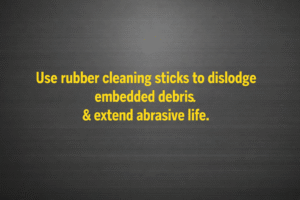 Minimalist vector graphic with gunmetal background and bright yellow text reading “Use rubber cleaning sticks to dislodge embedded debris & extend abrasive life.” 